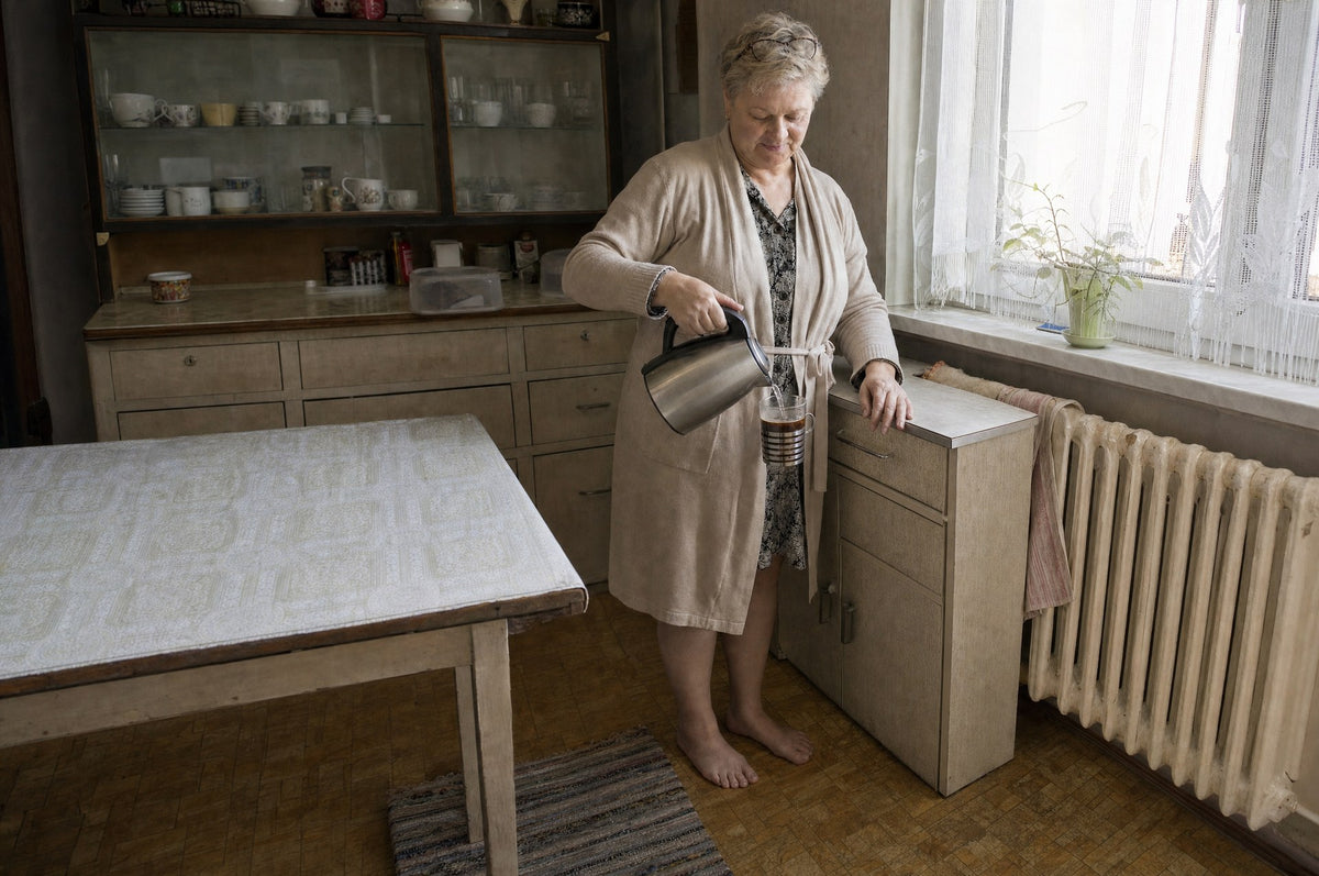 Pani Halina stoi boso na płytkach w kuchni rano i robi kawę